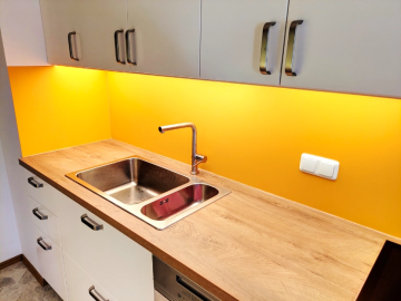 Moderne Küche mit gelb abgesetzten Wandpaneel als Rückwand, heller Holzarbeitsplatte und hellen Oberschränken.