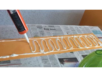 Mit einer Kartuschenpistole wird weißer Kleber in Schlangenlinien auf eine orangefarbene Wandschutzplatte aufgetragen, die auf mit Zeitungspapier abgedecktem Tisch liegt.